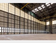 Große, leere Industriehalle mit hohen Decken und einer Wand aus Metallpaneelen, die von einer Reihe rechteckiger Fenster und Oberlichter durchzogen ist. Stahlträger und Beleuchtungskörper sind an der Decke sichtbar, während der Boden aus glattem Beton besteht.