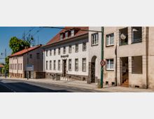 Straßenansicht eines Gebäudes mit der Aufschrift „Amtsgericht“ in einem hellen, zweigeschossigen Bau mit roten Ziegeldach und zahlreichen Fenstern. Daneben befinden sich mehrstöckige Wohnhäuser, ein Gehweg, Straßenlaternen und ein Verkehrsschild mit einer Geschwindigkeitsbegrenzung von 30 km/h.