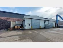 Industriegebäude mit Wellblechfassade und großen grauen Toren auf einem gepflasterten Gelände; daneben befinden sich Metallcontainer und gestapelte Paletten. Im Hintergrund ist ein Schutthaufen sowie ein blauer Kranarm zu sehen.