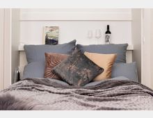 Ein gemütliches Schlafzimmer mit einem zentral platzierten Bett, das mit grauer Bettwäsche, dekorativen Kissen in verschiedenen Farben und Mustern sowie einer grauen Decke ausgestattet ist. Im Hintergrund stehen eine Flasche Wein, zwei Weingläser und ein kleines Landschaftsbild auf einem weißen Regal.