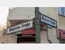 Straßenschilder markieren die Kreuzung von "Biebricher Straße" und "Mainzer Straße" vor einer hellen Putzfassade. Im Hintergrund sind ein Café-Schild und ein Teil eines Gebäudes sichtbar.