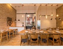 Restaurant-Innenraum mit Holzböden, freiliegenden Backsteinwänden und modernen Hängelampen. Zentral stehen lange Holztafeln mit gedeckten Plätzen, im Hintergrund eine Theke, ein Getränkekühlschrank und eine Glasschiebetür zur Küche.