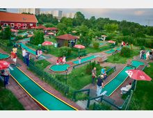 Eine Minigolfanlage im Freien mit mehreren Bahnen aus grünem Kunstrasen, umgeben von gepflegten Rasenflächen, Bäumen und roten Sonnenschirmen. Im Hintergrund ist ein rotes Gebäude mit Schrägdach sowie Wohngebäude sichtbar.