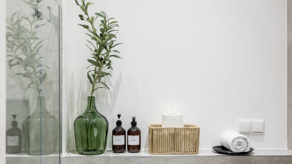 Modernes Badezimmerdetail mit Glasduschwand und grauen Fliesen; auf einer Ablage stehen eine große grüne Glasvase mit Zweig, zwei Spenderflaschen, ein geflochtener Korb mit Toilettenpapier und ein aufgerolltes Handtuch auf einer Schale vor weißer Wand mit Lichtschaltern.