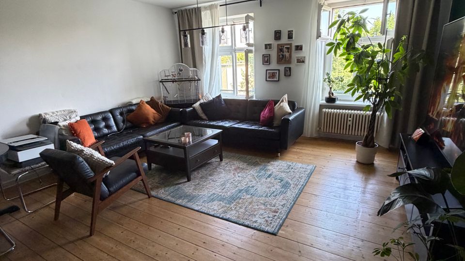 Helles Wohnzimmer mit breitem Holzdielenboden, schwarzem Ecksofa und Sessel um einen niedrigen Couchtisch auf einem Teppich. Zwei große Fenster mit Vorhängen, ein Heizkörper darunter, mehrere Zimmerpflanzen sowie eine Wand mit gerahmten Fotos prägen den Raum.