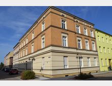 Ecke eines mehrstöckigen Wohngebäudes aus hellem Putz und orangefarbenem Backstein, mit hohen Fenstern und verzierten Fensterrahmen. Die Straße ist gepflastert, gesäumt von geparkten Autos und Straßenlaternen, mit angrenzenden ähnlich gestalteten Gebäuden.