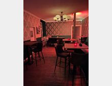 Ein gemütlicher Raum mit dunklem Holzboden, stilvoller Tapete mit Ornamentmuster und einer Deckenleuchte im Retro-Stil. Es gibt mehrere hohe Tische mit gepolsterten Barhockern, rote Kerzenlichter auf den Tischen sorgen für atmosphärische Beleuchtung.