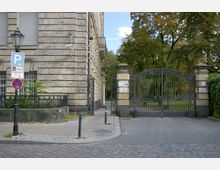 Steinernes Gebäude mit Fenstern und Rollläden an einer Straßenecke, daneben ein schmiedeeisernes Tor, das zu einem parkähnlichen Bereich mit Bäumen führt. Verkehrsschilder und Lampenpfosten befinden sich entlang des Gehwegs auf Kopfsteinpflaster.