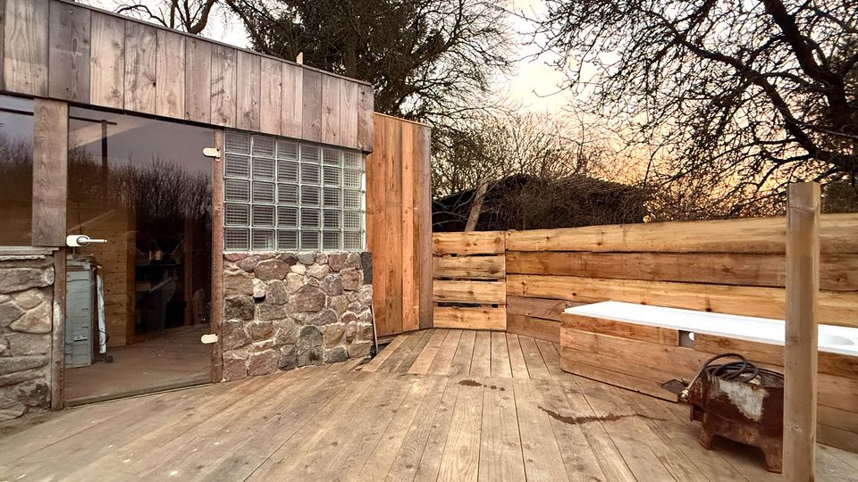 Holzterrasse im Außenbereich neben einem kleinen Gebäude mit Holzfassade, Steinsockel, Glastür und einem Fenster aus Glasbausteinen. Rechts steht eine lange, weiß verkleidete Bank vor einem hohen Holzsichtschutz; im Hintergrund sind kahle Bäume zu sehen.