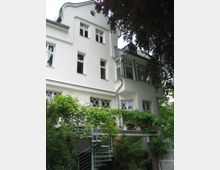 Weiße Fassade eines mehrstöckigen Wohnhauses mit großen Fenstern, Balkongittern im Jugendstil und einer Metallwendeltreppe, die von einem Balkon mit Pflanzenbewuchs herabführt. Vor dem Gebäude sind Bäume und Sträucher zu sehen.