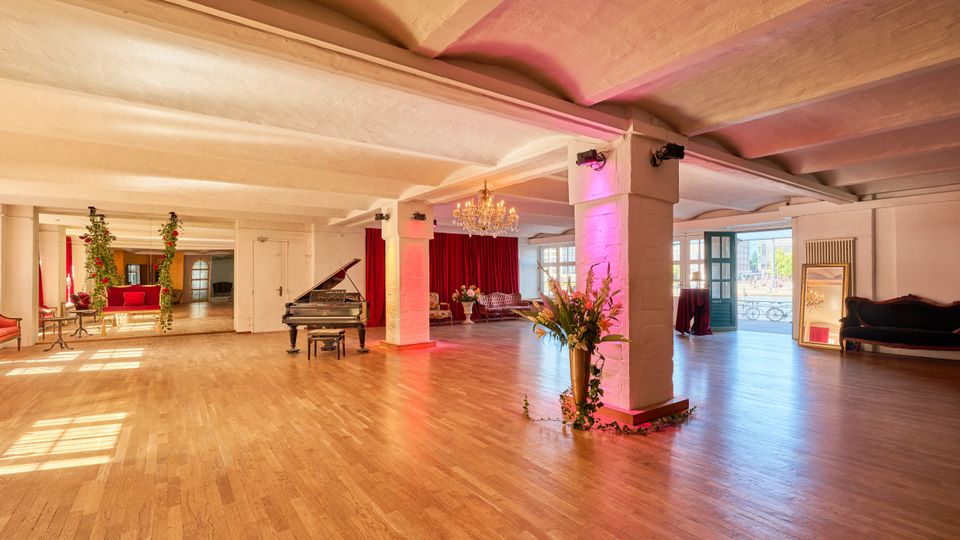 Großer, offener Veranstaltungsraum mit Holzparkett, niedriger Decke und weißen Stützpfeilern mit farbiger Beleuchtung. Im Raum stehen ein Flügel, ein Kronleuchter, rote Vorhänge sowie Sitzmöbel; große Fenster und eine geöffnete Tür lassen Tageslicht herein.