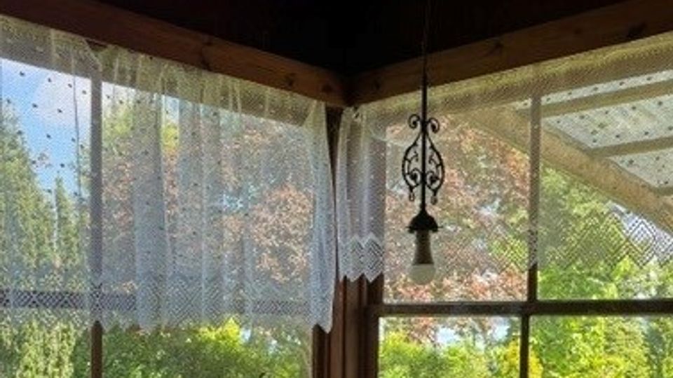 Blick aus einer verglasten Veranda mit Holzrahmen und dunkler Decke, an deren Ecke ein hängender Zieranhänger befestigt ist. An den Fenstern hängen transparente Spitzengardinen; draußen sind eine sonnige Rasenfläche und Bäume zu sehen.