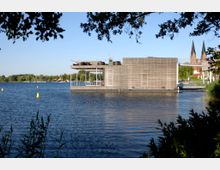 Ein modernes, quadratisches Gebäude mit Holzverkleidung steht auf einem See und ist über einen Steg verbunden; im Hintergrund sind Bäume und die Türme einer Kirche sichtbar. Gelbe Bojen schwimmen im Wasser, und der Vordergrund ist von Zweigen und Blättern eingerahmt.