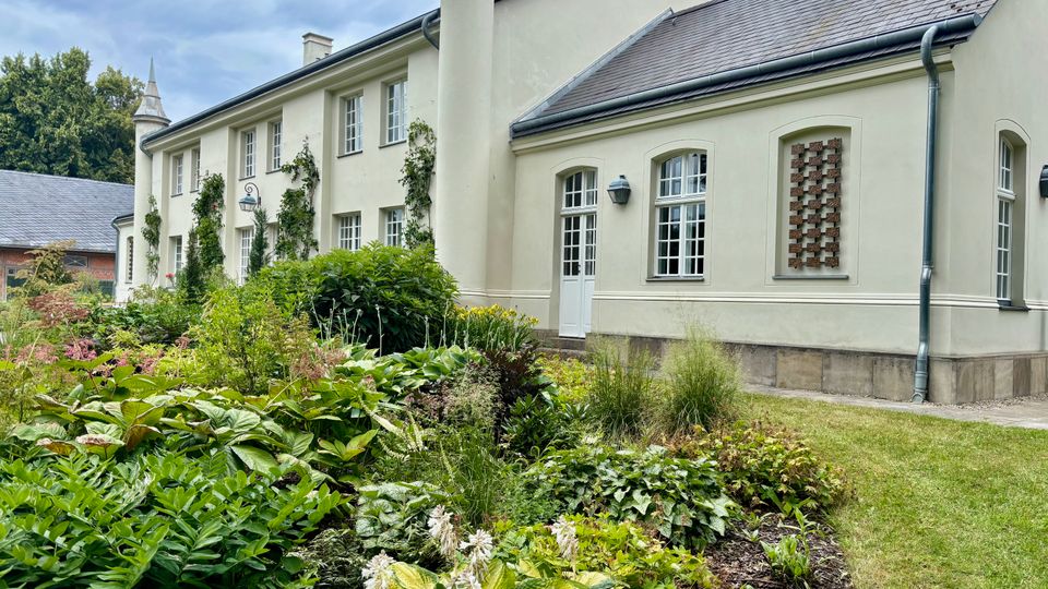 Hellfarbenes, zweigeschossiges Landhaus mit Satteldach, mehreren Kaminen, weißen Sprossenfenstern und einem runden Eckturm; an der Fassade wachsen Kletterpflanzen. Davor liegt ein gepflegtes Gartenbeet mit dichtem Grün und Stauden neben einem Rasenstreifen.