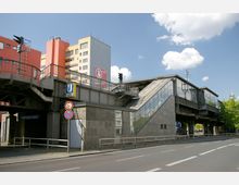 Bahnhof „U-Bhf Nollendorfplatz“ in Berlin mit erhöhter Bahnsteigstruktur, Stahl- und Glasarchitektur sowie einer großen Außentreppe. Im Hintergrund befinden sich bunte Wohngebäude und ein blauer Himmel mit wenigen Wolken.