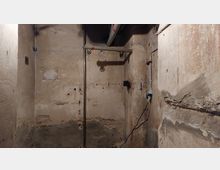 Ein leerer, ungepflegter Kellerraum mit bröckelnden Wänden und sichtbaren Rohrleitungen an der Decke sowie einer Steckdose mit Kabel an der rechten Wand. Der Boden und die Wandflächen zeigen Zeichen von Abnutzung und Feuchtigkeit.