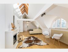 Helles Wohnzimmer in einem Dachgeschoss mit schrägen weißen Wänden und Holzbalken an der Decke. Sichtbar sind ein Kamin, ein Kuhfell-Teppich auf einem Holzfußboden, moderne Möbel wie Sessel und Sofa sowie ein halbrundes Fenster mit Blick nach draußen.