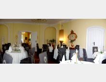 Eleganter Speiseraum mit hellgelben Wänden, weißen gewölbten Türen und dekorativen Zierelementen. Mehrere runde Tische sind mit weißen Tischdecken und gefalteten Stoffservietten gedeckt, umgeben von dunkel gepolsterten Stühlen; ein Kronleuchter hängt von der Decke, und ein Spiegel mit goldener Verzierung ist an der Wand angebracht.