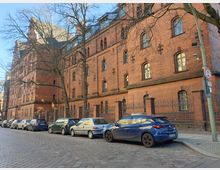 Straßenansicht eines mehrstöckigen, historischen Backsteingebäudes mit hohen, bogenförmigen Fenstern und einem spitz zulaufenden Dach. Im Vordergrund sind parkende Autos entlang einer gepflasterten Straße und ein Zaun mit Metallspitzen zu sehen, umgeben von kahlen Bäumen.