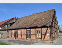 Fachwerkhaus aus rotem Ziegelstein mit dunklen Holzbalken und einem großen, mit Dachziegeln gedeckten Satteldach. Das Gebäude hat mehrere kleine Fenster und eine Holztür, es steht an einer gepflasterten Straße unter blauem Himmel.