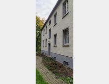 Mehrfamilienhaus mit heller Putzfassade und mehreren rechteckigen Fenstern, umgeben von einem kleinen, gepflasterten Weg und Beeten mit zurückgeschnittenem Grün. Im Hintergrund sind Bäume zu erkennen.