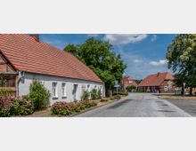 Straßenszene in einem Dorf mit traditionellen Häusern im Fachwerkstil und roten Ziegeldächern, umgeben von Blumenbeeten und Bäumen. Im Hintergrund ist eine Kreuzung zu sehen, umgeben von ländlicher Bebauung und einem klaren Himmel.