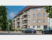 Mehrfamilienhaus mit vier Etagen und markanten Balkonen an einer Straßenecke, daneben Straßenschilder und parkende Autos. Im Vordergrund Gehwege mit Bäumen und Laternen, im Hintergrund Grünflächen und blauer Himmel.