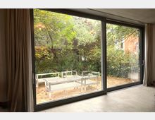 Großes Fenster mit Glastüren, das auf eine Terrasse mit weißem Gartenmobiliar und einen Garten mit Bäumen, Sträuchern und Pflanzen blickt; im Hintergrund ist eine Backsteinfassade eines Gebäudes zu sehen.