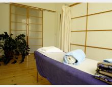 Minimalistisch eingerichteter Massageraum mit einem Massagebett, auf dem Handtücher und eine Nackenrolle liegen. Im Hintergrund sind helle Schiebetüren mit Holzrahmen, ein cremefarbener Vorhang und eine Topfpflanze sichtbar.