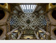Blick auf eine kunstvoll ornamentierte, gewölbte Decke mit geometrischem Muster in einer historischen Innenraumarchitektur. Sichtbar sind auch filigrane Verzierungen aus Stein und ein großes Oberlicht mit Glasfenstern, das den Raum erhellt.