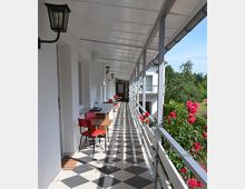 Überdachter Außengang mit diagonalen schwarz-weißen Fliesen, weißen Wänden, roten Stühlen an Tischen und blühenden Rosen entlang eines weißen Geländers; im Hintergrund sind Bäume und blauer Himmel zu sehen.