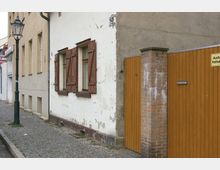 Straßenszene mit Kopfsteinpflaster, einer Hauswand mit abgenutztem Putz und Holzfensterläden sowie einer angrenzenden Holztorwand mit gemauertem Pfeiler und einem Schild mit der Aufschrift „Ausfahrt freihalten“. Links steht eine traditionelle schwarze Straßenlaterne.