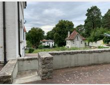 Hofbereich mit einer niedrigen Steinmauer und einer kleinen Treppe im Vordergrund, flankiert von weißen Gebäuden mit roten Ziegeldächern. Im Hintergrund Bäume und weitere Häuser in einer grünen, ländlichen Umgebung.