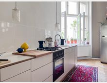 Helle, moderne Küche mit weißer Einbauzeile, Gasherd und Spüle unter einem großen Fenster mit geöffneten Flügeln. Auf der Arbeitsfläche stehen Küchenutensilien, Pflanzen und Dekoobjekte; ein roter Teppich bedeckt den Holzboden.