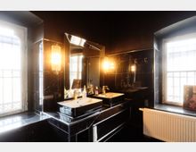 Modernes Badezimmer mit schwarzen, marmorierten Wand- und Bodenfliesen, Doppelwaschbecken und großem Spiegel mit warmen Wandleuchten. Zwei große Fenster lassen Tageslicht herein; darunter befinden sich eine breite Fensterbank und ein Heizkörper.