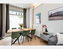 Heller Essbereich in einem modernen Wohnraum mit großem Fenster, dunklen Vorhängen und drei hängenden Pendelleuchten über einem Holztisch. Grüne Polsterstühle stehen um den Tisch; an der Wand hängen gerahmte Bilder neben einem Heizkörper, daneben ein kleines Sideboard und ein Teil eines grauen Sofas.