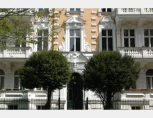 Fassade eines historischen Wohnhauses mit gelbem Klinkermauerwerk, weißem Stuck und verzierten Fensterelementen. Vor dem Gebäude befinden sich zwei dicht belaubte Bäume und ein schmiedeeiserner Zaun.