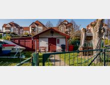 Kleines Gebäude mit rotem Dach und weißer Fassade, beschriftet mit „Angler Sektion Wildau“, umgeben von einem kleinen umzäunten Garten. Im Vordergrund ein Weg, ein Boot auf einem Anhänger und beschnittene Baumstämme, im Hintergrund mehrere mehrstöckige Häuser mit roten Ziegeldächern.