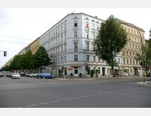 Ecke eines belebten Straßenzuges mit mehrstöckigen Altbaugebäuden, verziert mit Fassadendekorationen und großen Fenstern; im Erdgeschoss befinden sich Geschäfte und Restaurants. Die Straße ist umgeben von Bäumen sowie parkenden Autos und umfasst Fußgängerüberwege und Ampeln.