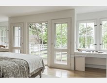 Helles Schlafzimmer mit Holzboden, großem Bett und weißer Bettwäsche, umgeben von mehreren Fenstern und einer Balkontür, die auf einen Balkon mit Blick auf Bäume und ein Haus mit Ziegeldach führen. Ein weißer Schreibtisch mit Dekorationsobjekten steht unter den Fenstern.