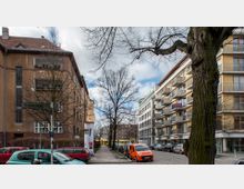 Straßenszene mit mehrstöckigen Wohngebäuden auf beiden Seiten, links ein älteres Gebäude mit hellbrauner Fassade, rechts ein modernes Gebäude mit Balkonen. Im Vordergrund stehen geparkte Autos und Bäume ohne Laub, im Hintergrund ist eine gelbe Straßenbahn sichtbar.