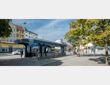 U-Bahnhof Franz-Neumann-Platz in Berlin mit überdachtem Eingangsbereich, umgeben von mehrstöckigen Wohnhäusern, Bäumen und Geschäften. Ein Flugzeug fliegt am klaren Himmel über die Szene, und Personen befinden sich auf dem Gehweg.