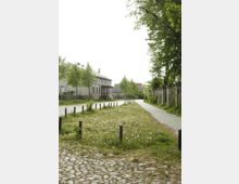 Gepflasterte Straße mit angrenzender Grünfläche, gesäumt von Pollern, links historische Gebäude mit Giebeldächern, rechts ein schmaler Kiesweg und eine Steinmauer, umgeben von hohen, grünen Bäumen.