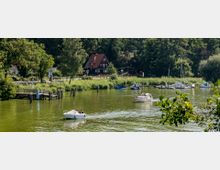 Flusslandschaft mit grünem Wasser, kleinen Motorbooten und Segelbooten, die an einem schmalen Steg anlegen. Im Hintergrund sind ein rotdachiges Haus, Bäume und dichte Vegetation zu sehen.