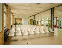 Großer, heller Veranstaltungsraum im Industrie-Loft-Stil mit Reihen weißer Stühle, hohen Fenstern und freiliegenden Backsteinwänden. Sichtbare Stahlträger, Akustikdeckenpaneele, Pflanzen sowie Sitzbereiche und Getränkeautomaten im Hintergrund.