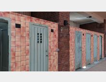 Reihe von kleinen Zellen mit rostigen Metalltüren in einem Gebäude, dessen Wände teils aus roten Ziegelsteinen und teils aus orangefarbenen Fliesen bestehen. Die Türen haben schmale Lüftungsschlitze und leicht abgenutzte Oberflächen.