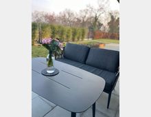 Ein moderner Außenbereich mit grauen Fliesen, einem schwarzen Metalltisch und einer dunkelgrauen Sitzbank. Auf dem Tisch steht eine Glasvase mit rosa Blumen, im Hintergrund sind eine Hecke, Bäume und ein Garten sichtbar.