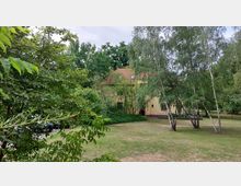 Gelbes Gebäude mit rotem Ziegeldach, umgeben von dichtem Grün und Bäumen, auf einer Fläche mit Rasen. Im Vordergrund stehen Birken und Sträucher; am linken Bildrand sind geparkte Autos sichtbar.