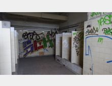 Ein verlassenes öffentliches Toilettenhaus mit beige gefliesten Wänden, beschmiert mit zahlreichen Graffiti in verschiedenen Farben. Die Kabinenwände sind teilweise beschädigt, und der Raum zeigt sichtbare Abnutzungsspuren.