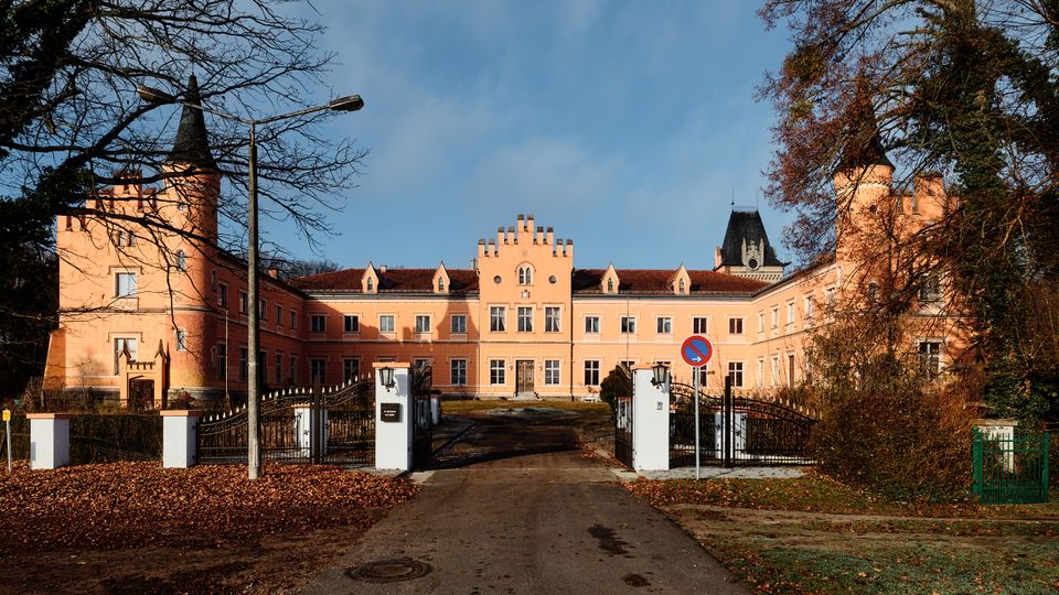 Frontansicht eines rosafarbenen Schloss- oder Herrenhauskomplexes mit zentralem Giebel und seitlichen Türmen, erreichbar über eine Zufahrt durch ein schmiedeeisernes Tor. Vor dem Gebäude stehen weiße Torpfeiler und Laternen, umgeben von Bäumen und herbstlichem Laub.